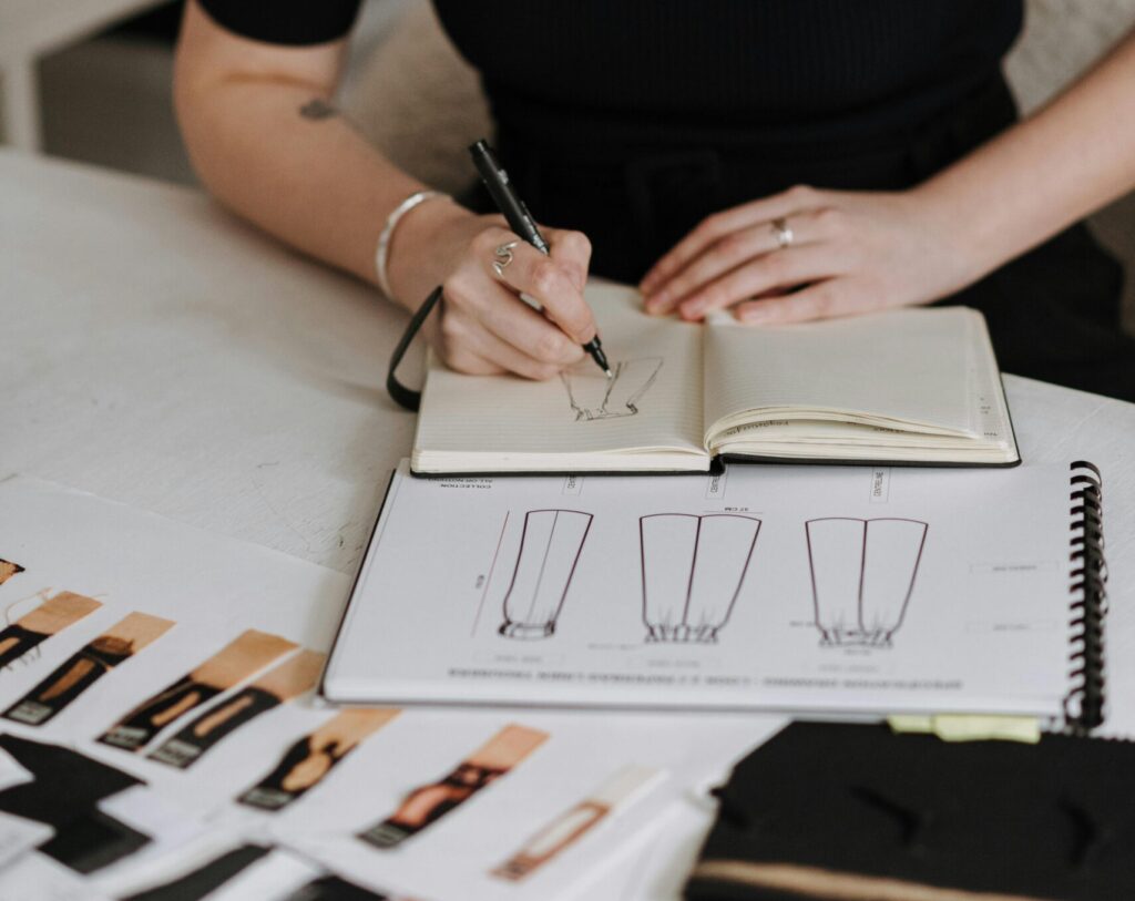 Anonymous female designer drawing sketch of new clothes while sitting at table with samples of fabrics during work in studio